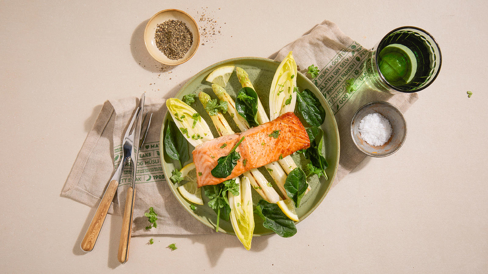Grillet laks med hvit asparges og spinat, servert med sikoriblader og sitronskiver på lys grønn asjett. Bestikk og grønt vannglass rundt.