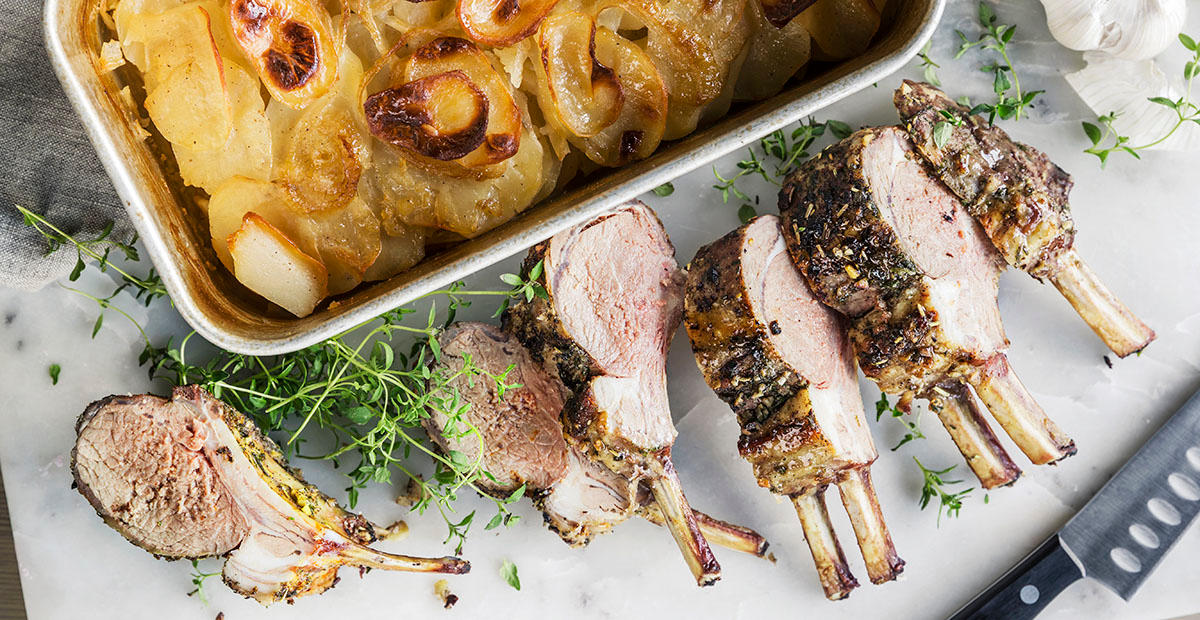 Lammecarré med urtelokk delt i skiver, og servert på hvit marmorfjøl. Potetform ved siden av, og ferske urter omkring. Kniv på siden.