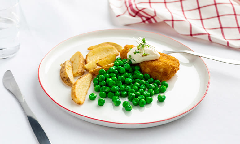 Panert torsk med potetbåter og erter, servert på en hvit tallerken med rød kant på hvitt bord med rødrutet kjøkkenhåndkle ved siden av. Torsken har en klatt aioli på toppen.