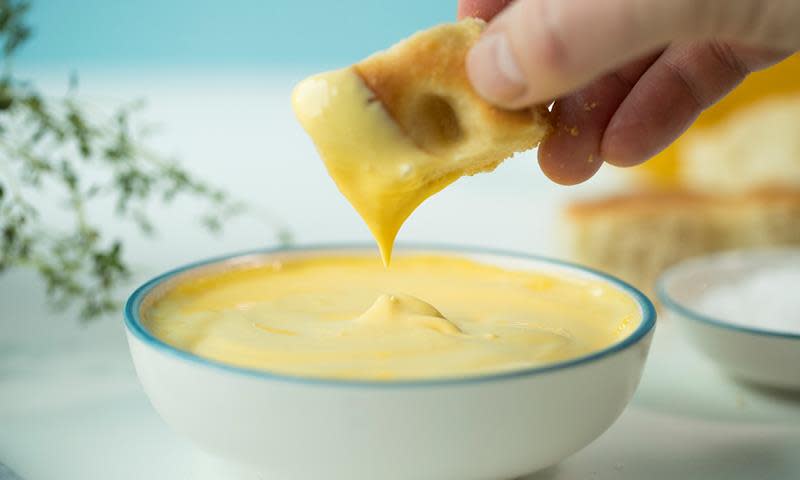 Rask hjemmelaget aioli i lys skål med blå kant. Hånd holder en bit focaccia og dypper i aiolien. Ferske urter i bakgrunnen.