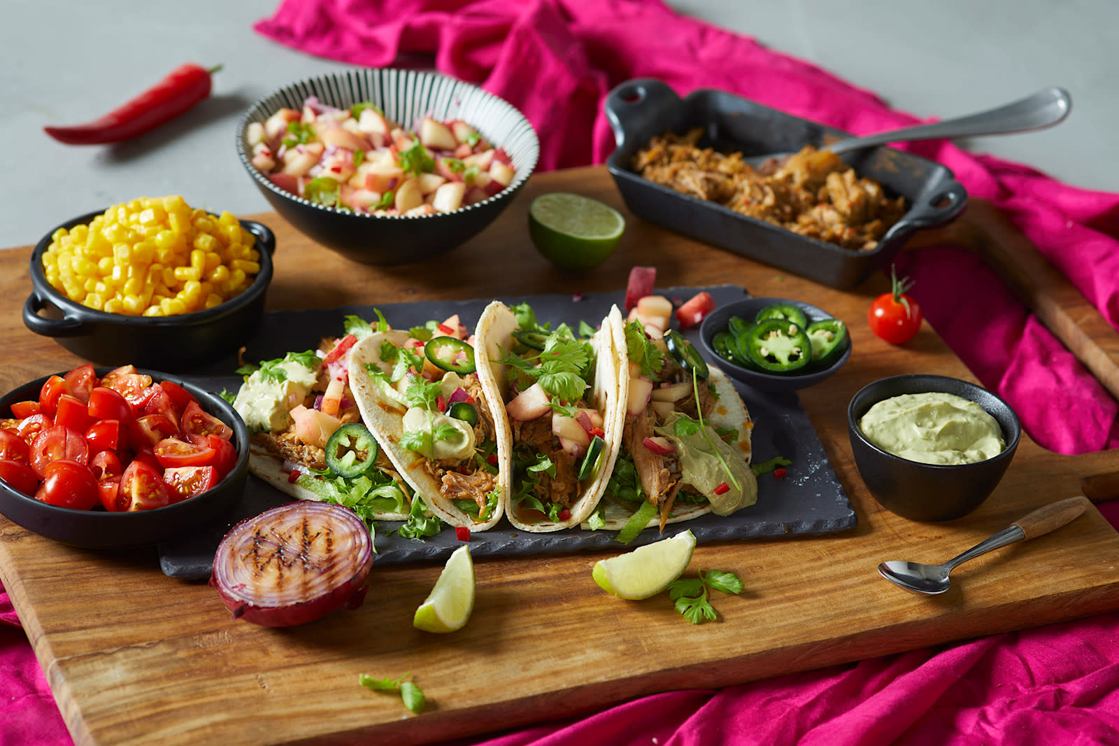 Pulled pork carnitas med ferskensalsa og avokado crema servert i små maislefser med flere skåler tilbehør rundt. Liten skål med pulled pork i bakgrunnen.