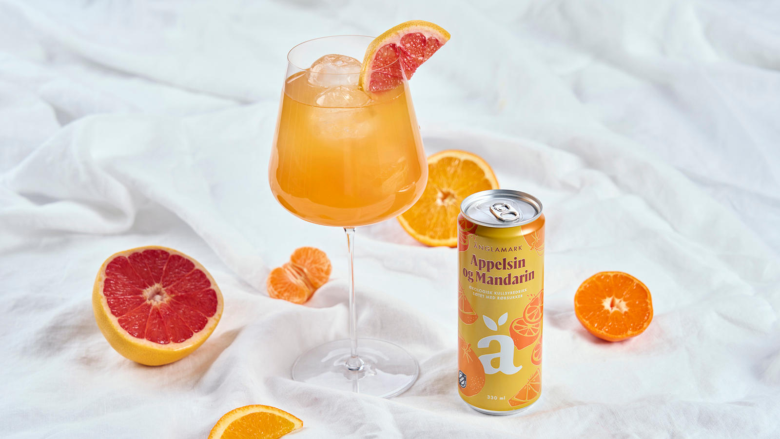 Änglamark Spritz mocktail servert i stort vinglass ved siden av en boks Änglamark Appelsin og Mandarin. Biter av frisk mandarin, appelsin og blodappelsin rundt.