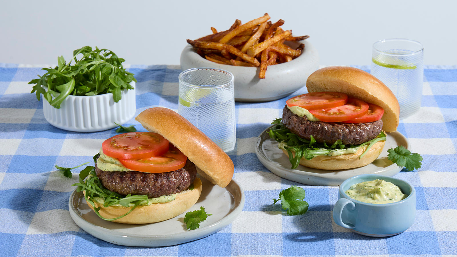 Hamburger med hjemmelaget korianderaioli og skinny fries servert på lysegrå asjetter på blårutet duk. Skål med skinnyfries og ruccola i bakgrunnen.