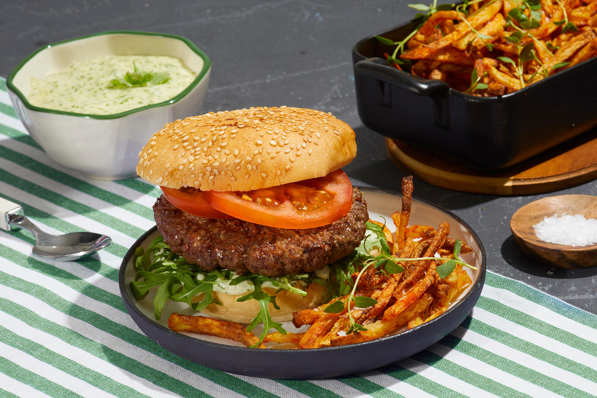 Hamburger med korianderaioli og skinny fries servert i hamburgerbrød med ruccola og skivet tomat, på blå asjett på grønnstripete duk. Skåler i bakgrunnen med grovt salt, korianderaioli og skinny fries med urter.
