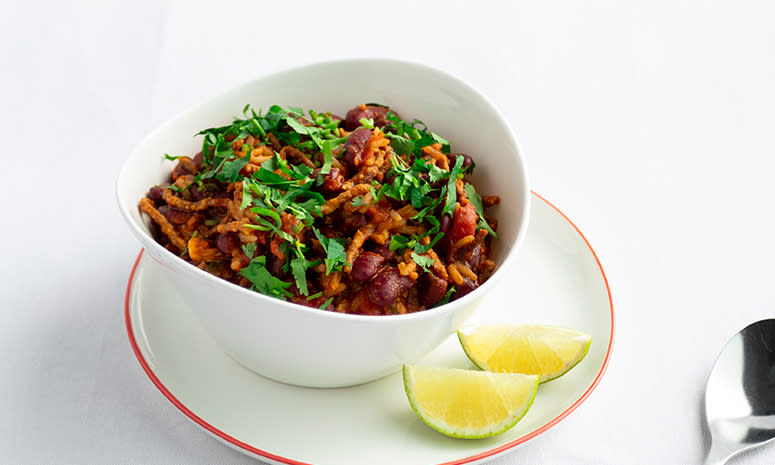 Chili sin carne i en dyp hvit skål, oppå en tallerken men rød kant, med to limebåter på høre siden