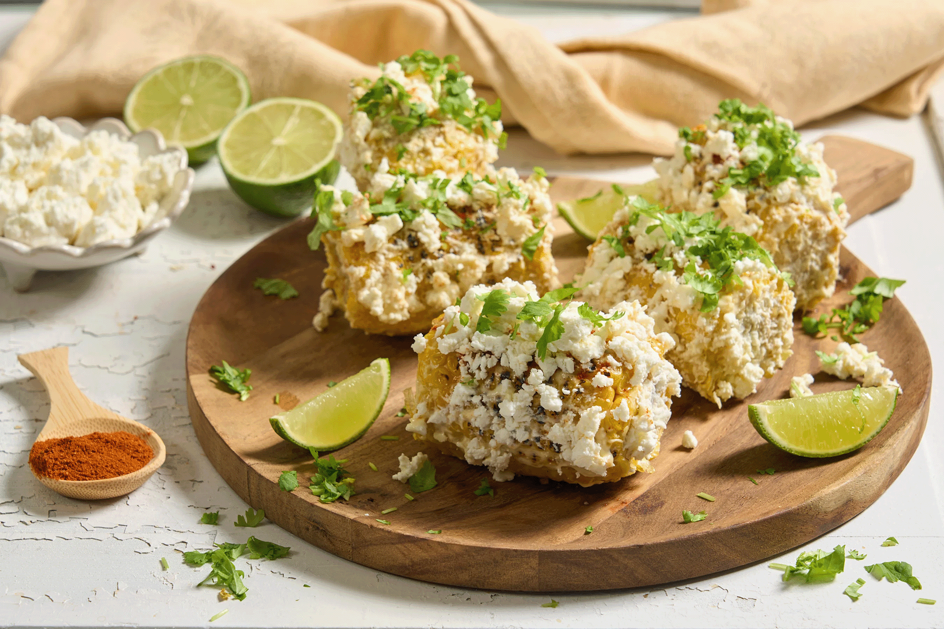 Elotes - Meksikansk street corn med fetaost og koriander, servert på rund trefjøl med limebåter. Halve lime, liten skål med fetaost og skje med chilipulver ved siden av.