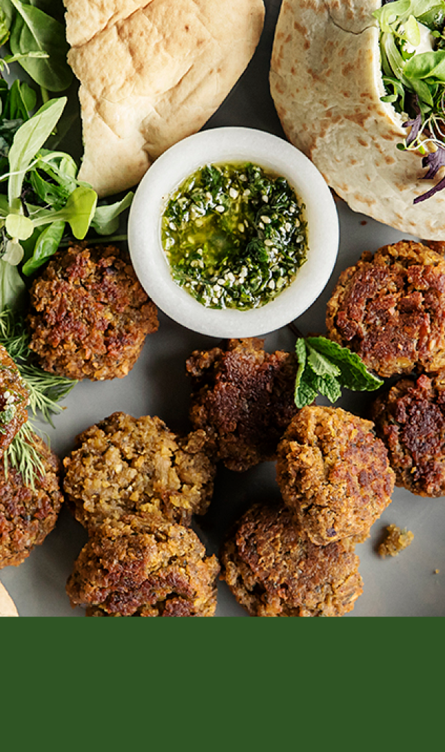 En hånd holder falafel over et falafelfat med grønt, dipp og pitabrød øverst og grønn bakgrunn nederst