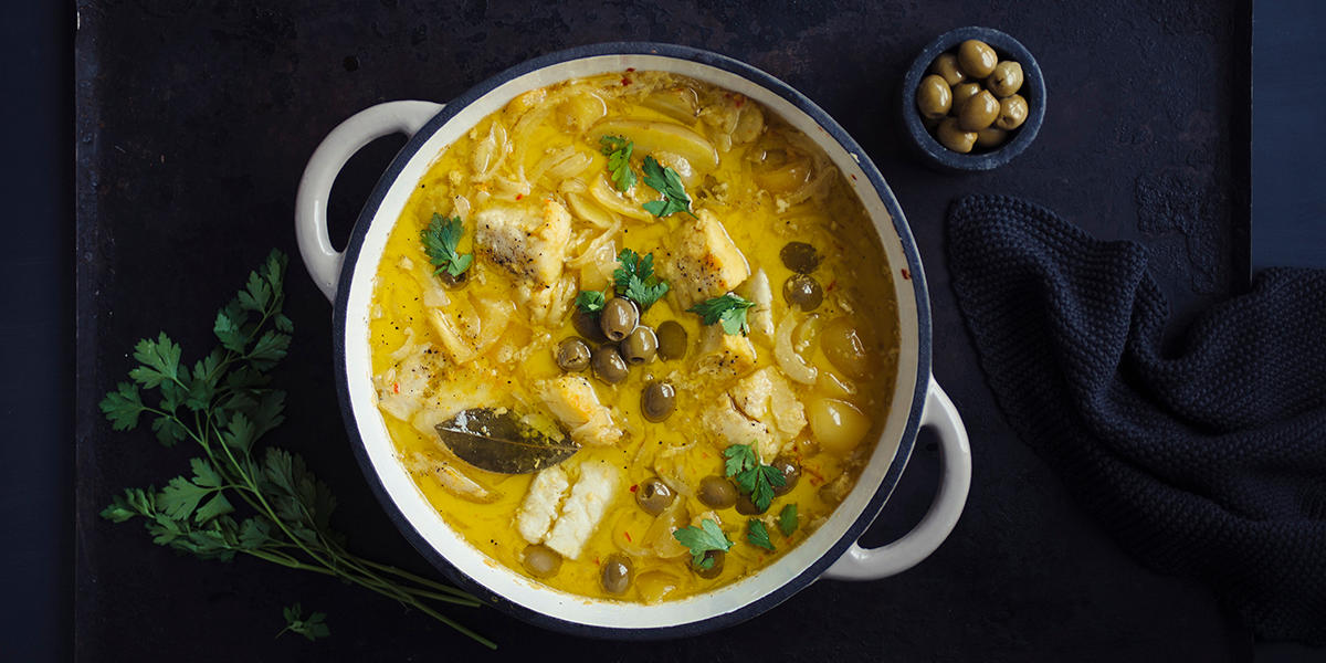 Hvit bacalao med safran, oliven og ferske urter servert i hvit støpejernskjele på mørk bakgrunn med ferske urter og en liten mørk skål med oliven ved siden av.