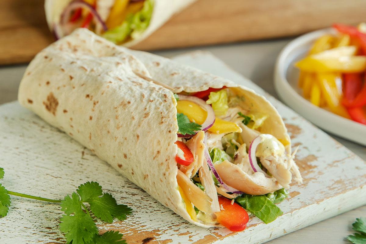 Kyllingwrap i tortillalefse, med grillet kylling, strimlet salat, rød paprika, mango, rødløk og karridressing. Servert på lysmalt trefjøl.