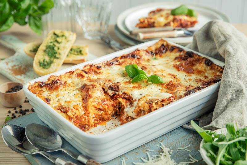 Klassisk lasagne med kjøttsaus og bechamelsaus, servert i hvit ildfast form med ferske urter og hvitløksbrød. Dekket middagsbord.