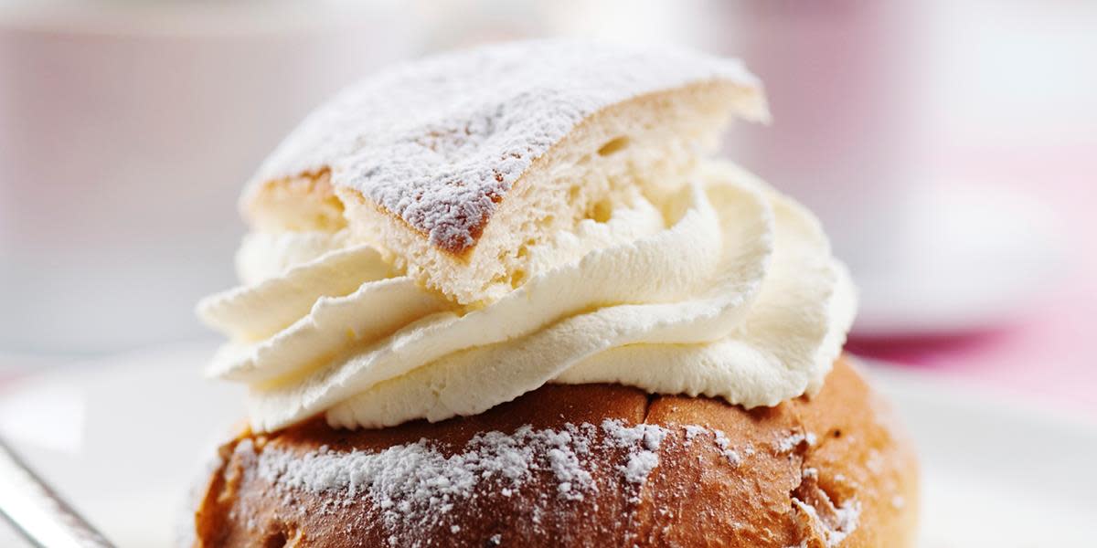 Svenske semlor; svenske fastelavnsboller fylt med mandelmasse og pisket krem. En bolle med toppen klippet av, fylt med krem med toppen som lokk, servert på hvit tallerken med rosa bakgrunn.