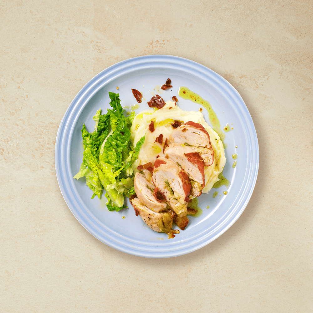 Fylt kyllingfilet med mozzarella og pesto, servert over potetmos, med hjertesalat på siden. Lys blå tallerken.