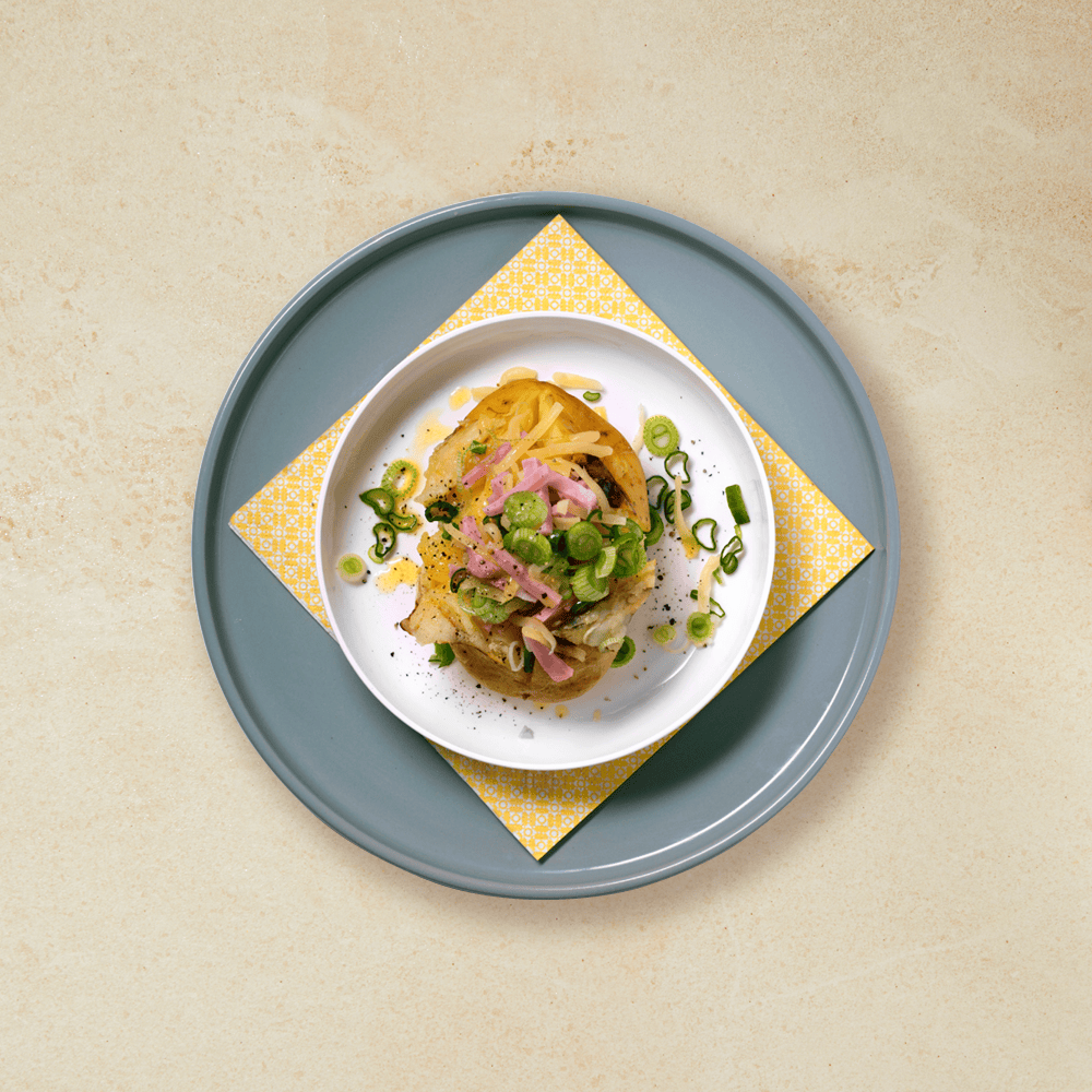 Bakt potet med ost og skinke servert på hvit skål på blå tallerken med gul serviett. Finsnittet vårløk strødd på toppen.