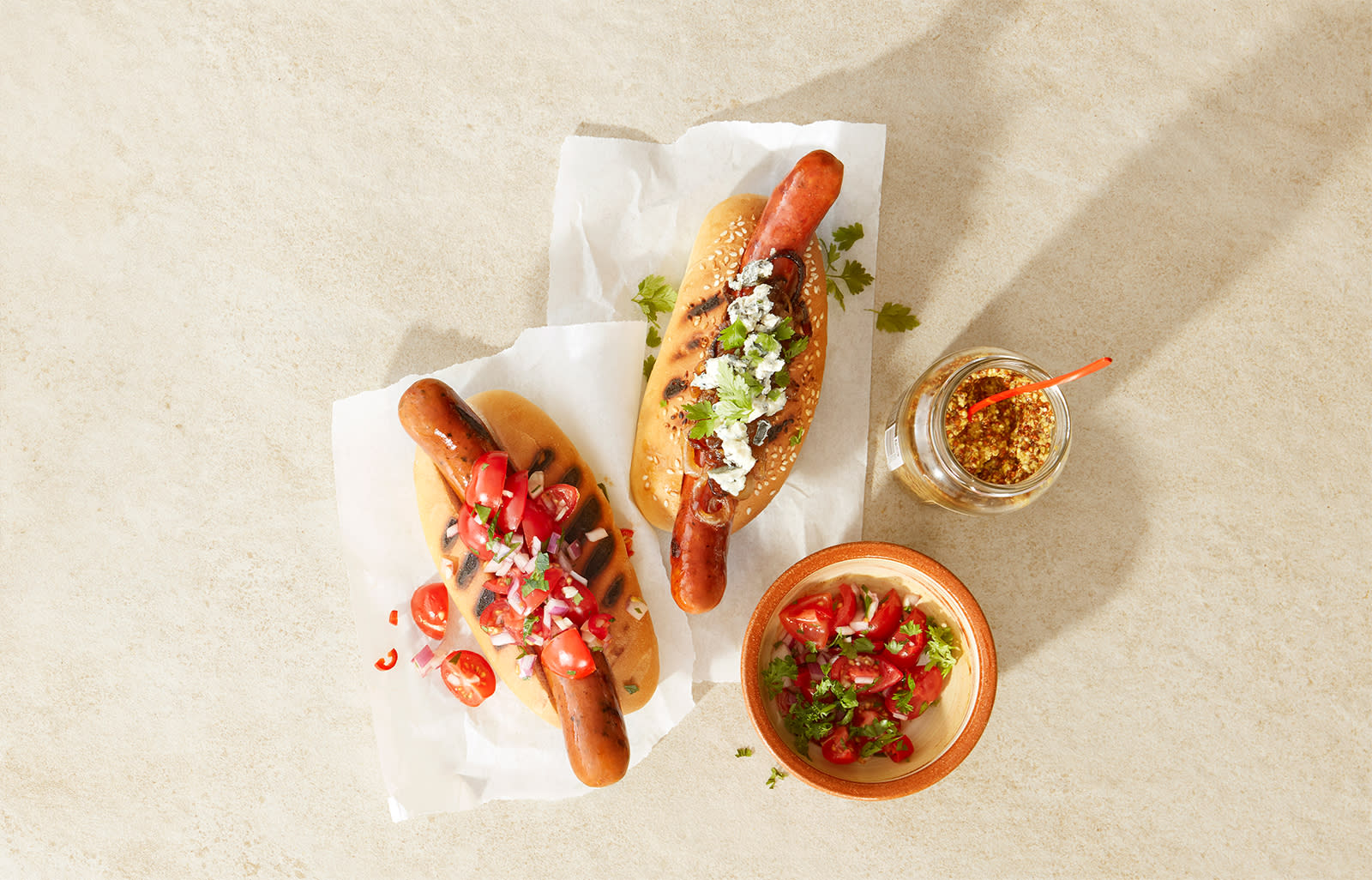 2 gode tilbehør til pølser; spicy relish og karamellisert løk med blåmuggost, servert på to grillede pølser i brød. To små skåler med spicy relish og sennep ved siden av.