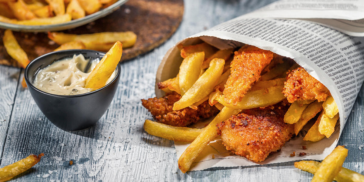 Hjemmelaget fish and chips servert på trebord i kremmerhus av avispapir. Liten mørk skål med remulade ved siden av, og skål med chips i bakgrunnen.