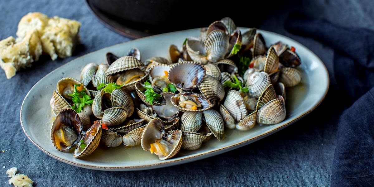 Hjerteskjell med hvitløk og persille på avlangt grått fat, servert på mørk linduk, med lyst brød i bakgrunnen.
