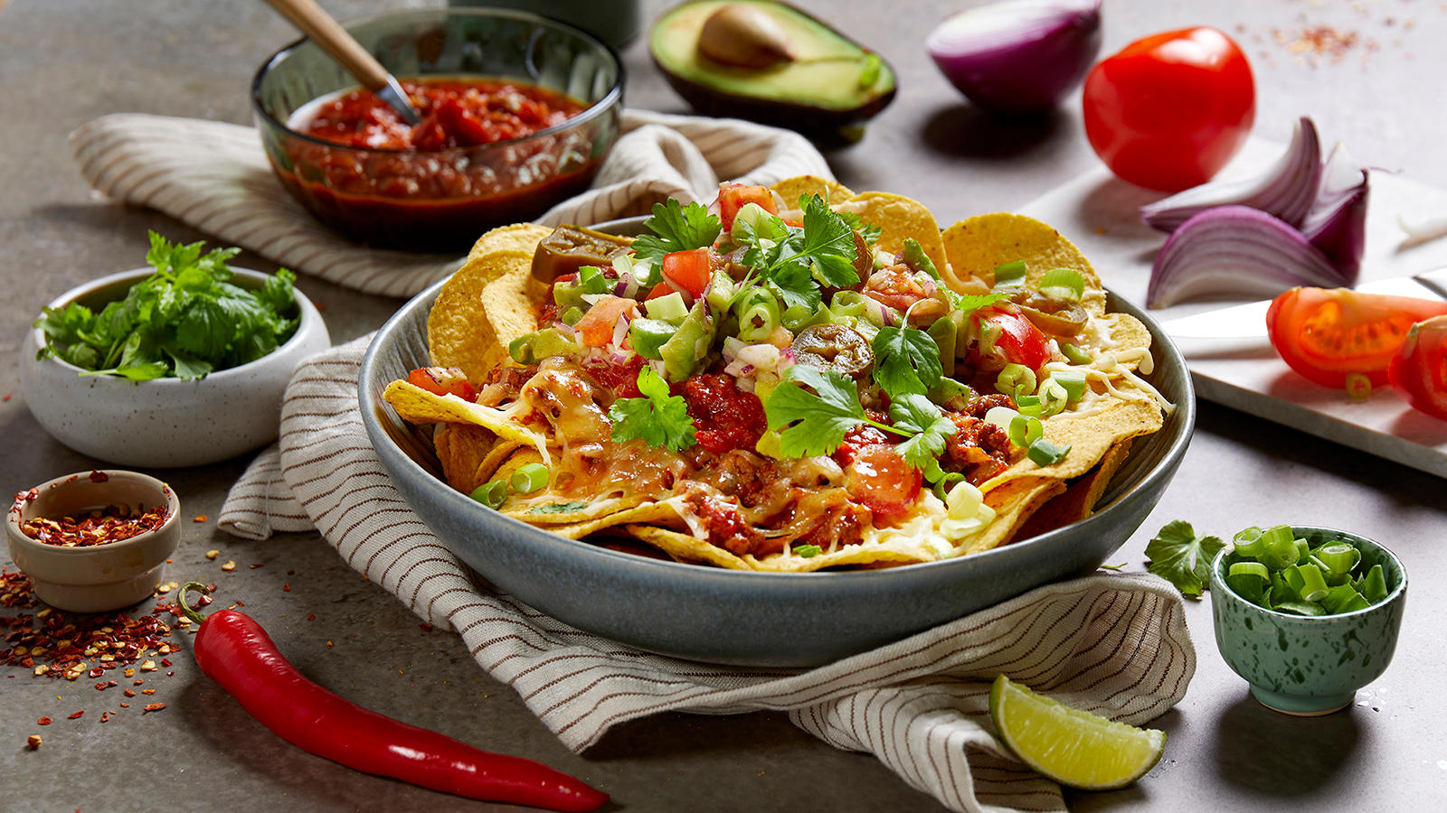 Nachos med kjøttdeig og smeltet ost servert med salsa, tomater, koriander, vårløk og jalapeños. Friske grønnsaker og nachotilbehør i små skåler rundt.