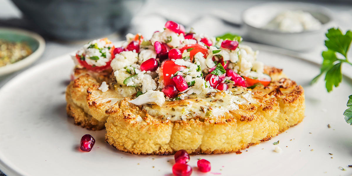Gratinert blomkålbiff med eksotisk tilbehør av krydret aioli, raspet blomkål, tomatbiter, granateplekjerner og friske urter servert på hvit tallerken.
