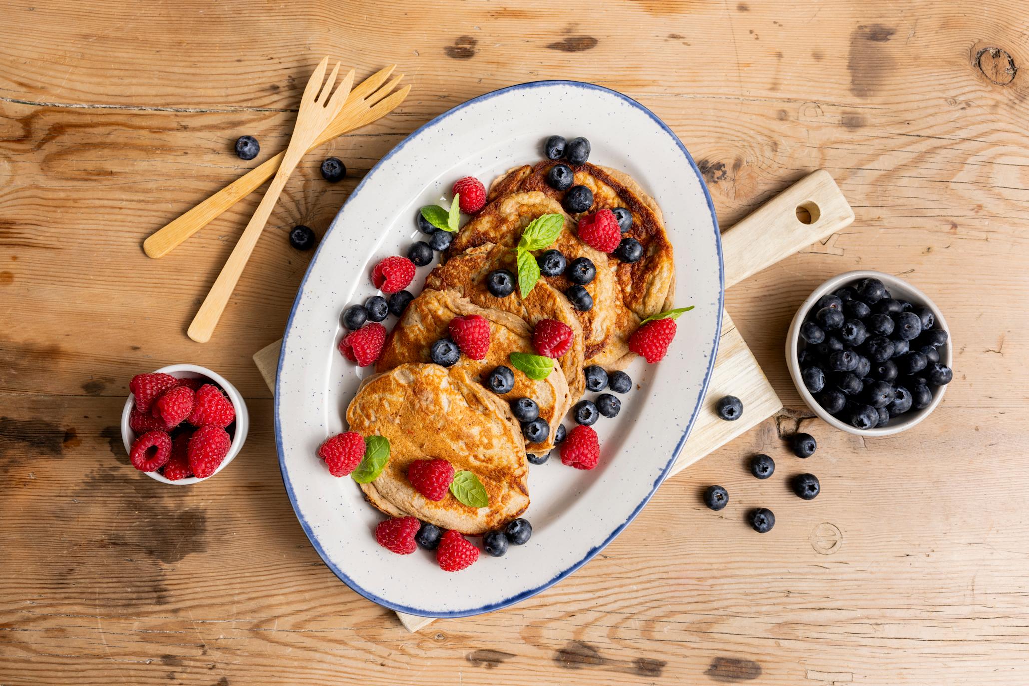 Sunne matpakkepannekaker på en oval tallerken på et trebord, dandert med bringebær, blåbær og mynte. Skåler med ferske bringebær og ferske blåbær ved siden av.