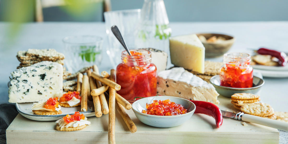 Ostefat med flere oster og et glass og en skål med chilimarmelade, brødpinner, kjeks og rød chili.