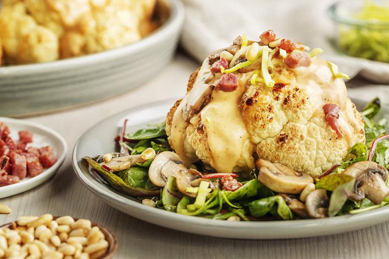 Gratinert blomkål med bacon og ostesaus, servert helt på grå asjett på en seng av purre og bacon. Skål med pinjekjerner ved siden av.