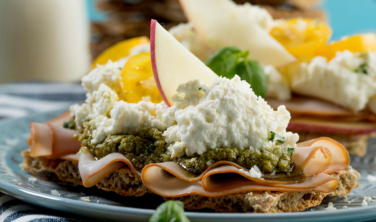 Hjemmelaget cottage cheese servert på grovt knekkebrød med skinke, pesto, gule tomater og epleskiver på en blå tallerken.