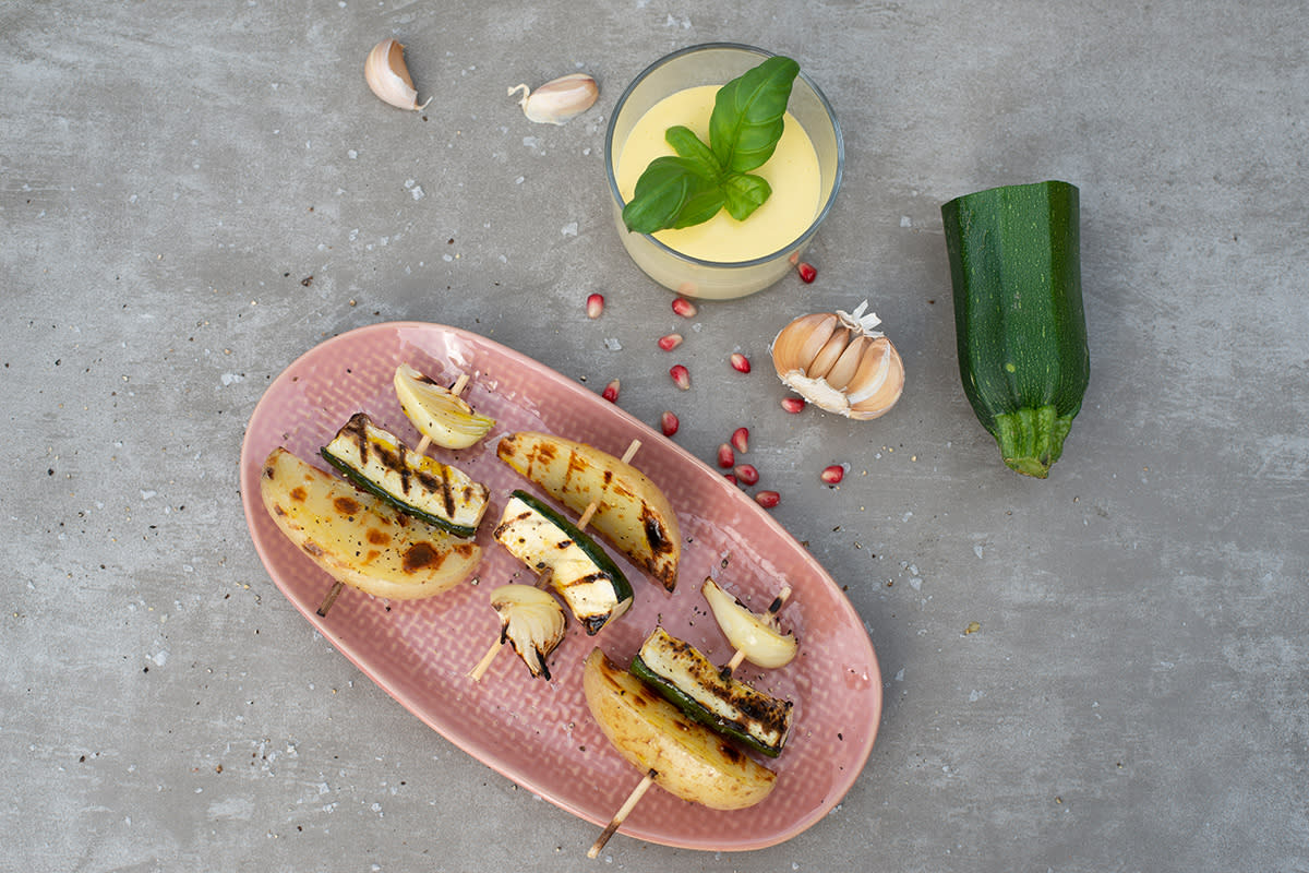 Tre grillspyd med småløk, squash og nypotet på en rosa tallerken ved siden av et glass aioli og grønnsaker.