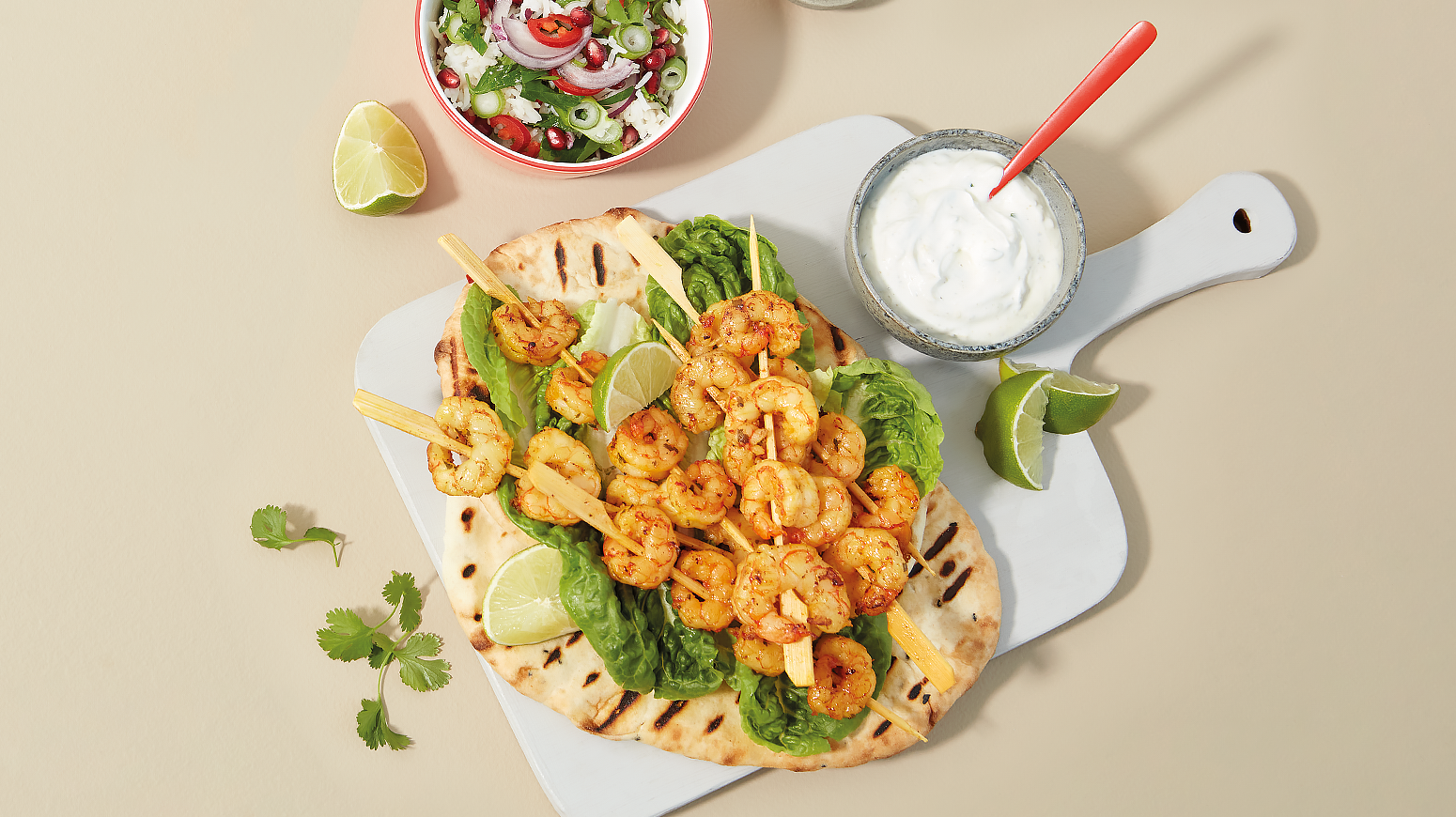 Indiske rekespyd servert på grillet nanbrød med salatblader og limebåter på hvit fjøl. Rød skål med rissalat og en mindre skål med raita ved siden av.