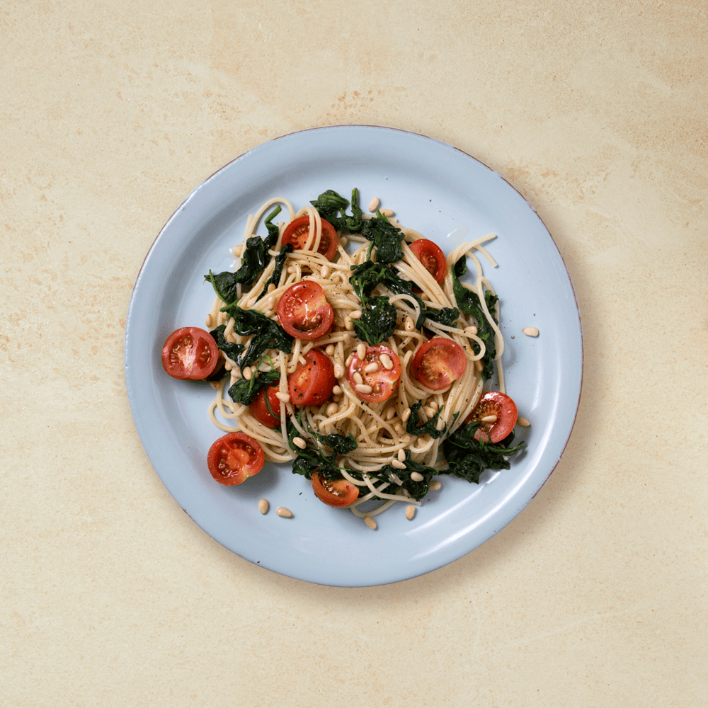 Vegetarpasta med spinat og tomat, overstrødd med pinjekjerner og servert på lys blå tallerken.