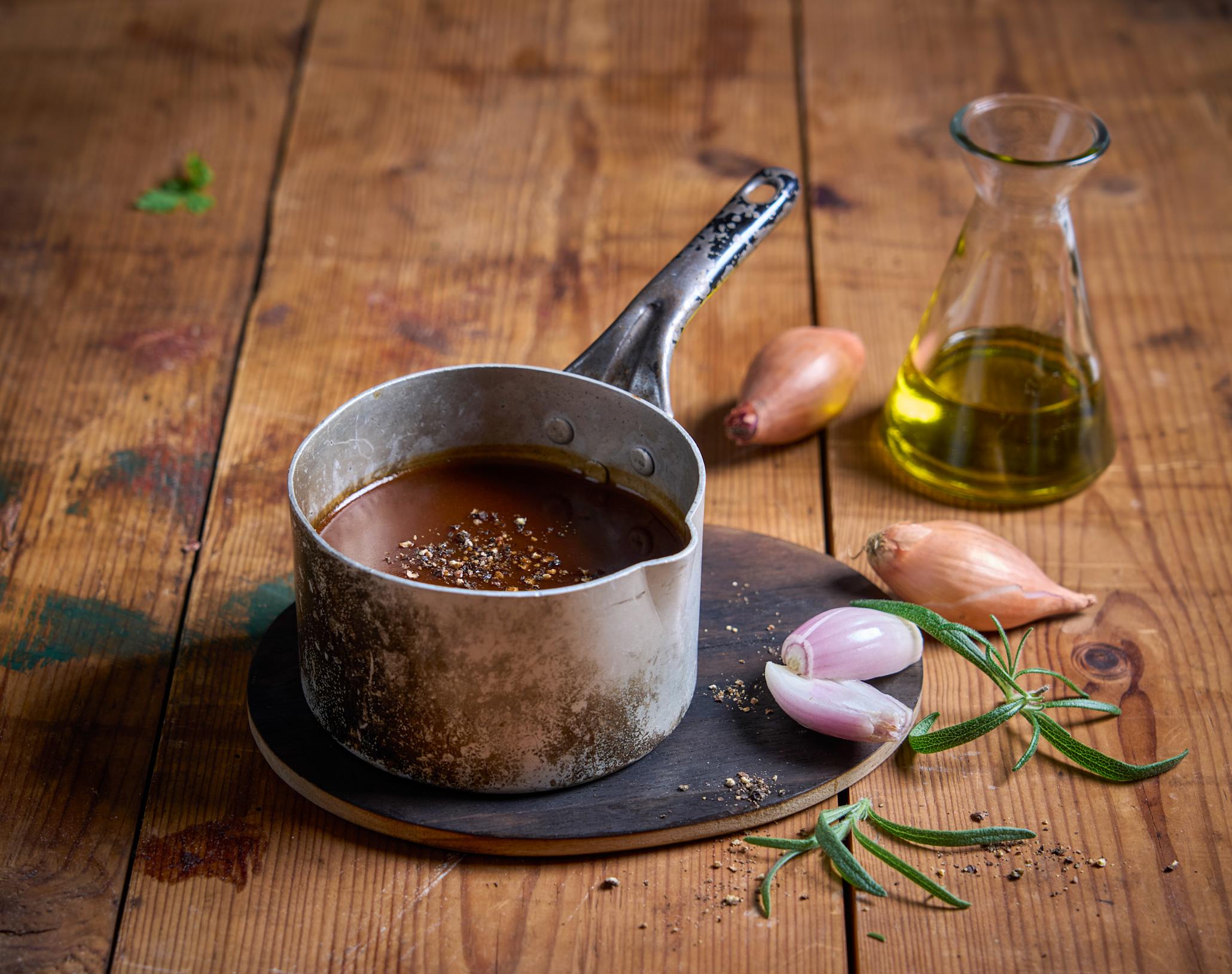 Rødvinssaus til skrei servert i liten grå metallkjele på rustikk trebord. Rundt kjelen er det strødd hel sjalottløk med og uten skall, og friske urter. En liten karaffel med olje står i bakgrunnen.