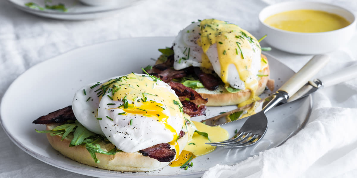 Eggs benedict - posjerte egg med Hollandaisesaus servert på en delt English muffin med bacon og salat, servert på en hvit tallerken på hvitt bord. Liten hvit skål med Hollandaise ved siden av.