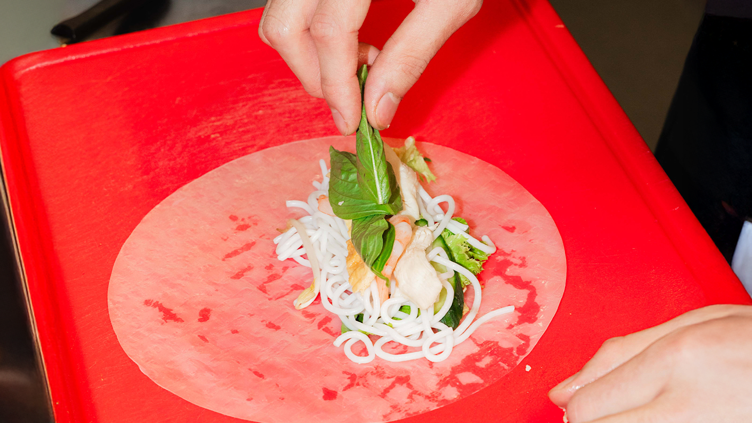 Nærbilde av hender som legger fersk basilikum og andre urter oppå risnudler og kylling i et rispapir, på en rød skjærefjøl, under lagingen av en vietnamesisk sommerrull.
