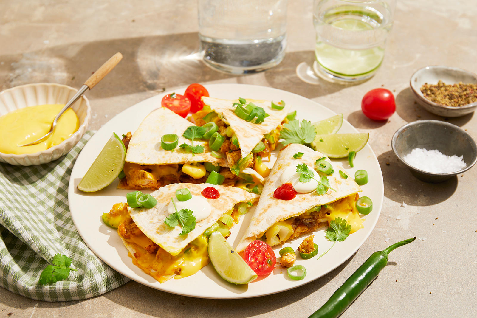 Quesadilla med kavli cheddar dipp og karamellisert løk, servert på hvit middagstallerken med små skåler dipp og krydder rundt. Quesadillaene er toppet med limebåter, cherrytomater, vårløk og koriander.