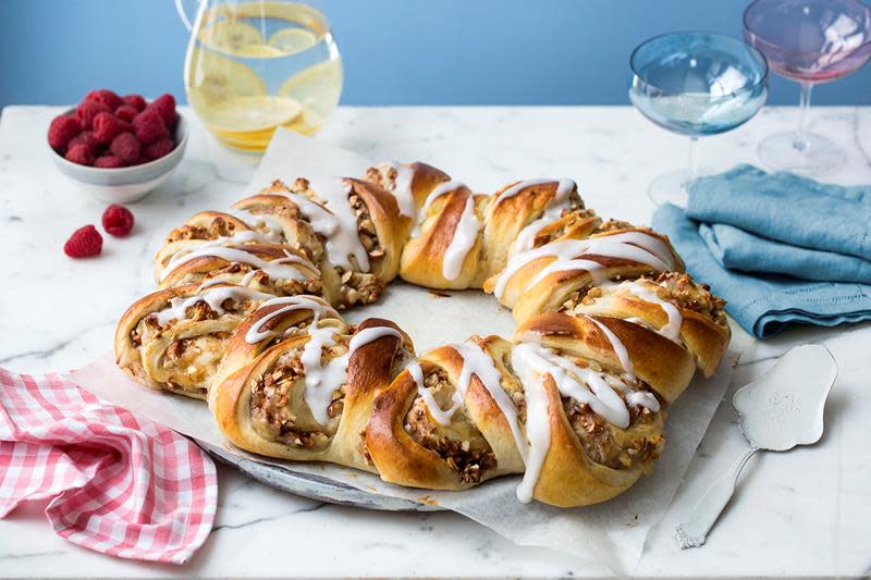 Fabelaktig klippekrans med eple og marsipan, servert med melisglasur på toppen, på hvitt marmorbord. Skål med ferske bringebær, og mugge med vann og sitronskiver i bakgrunnen.