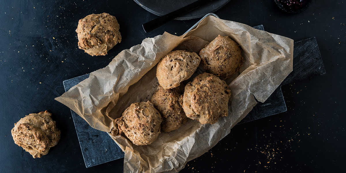 Kaldhevede grove rundstykker toppet med frø, servert i metallform med lysebrunt bakepapir på mørk steinfjøl. To rundstykker ligger på siden av formen.