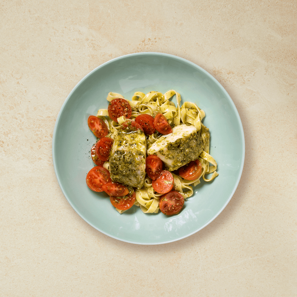 Ovnsbakt torsk med pesto, pasta og cherrytomater, i en grønn skål på beige bakgrunn.