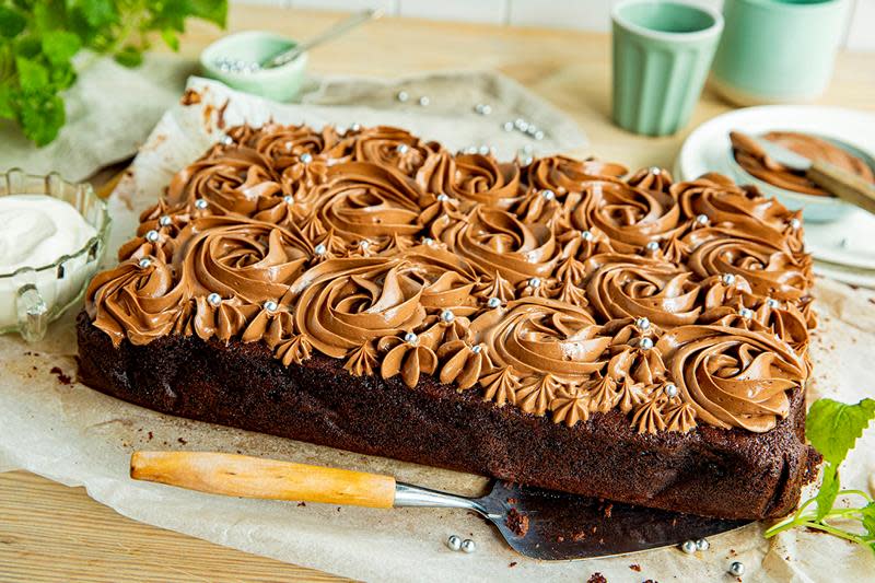 Saftig sjokoladekake laget i langpanne, pyntet med sjokoladesmørkrem i rosemønster med sølvkuler i sukker. Kaken er servert på lyst bakepapir på lyst trebord med en skål sjokoladekrem i bakgrunnen og friske urter ved siden av.