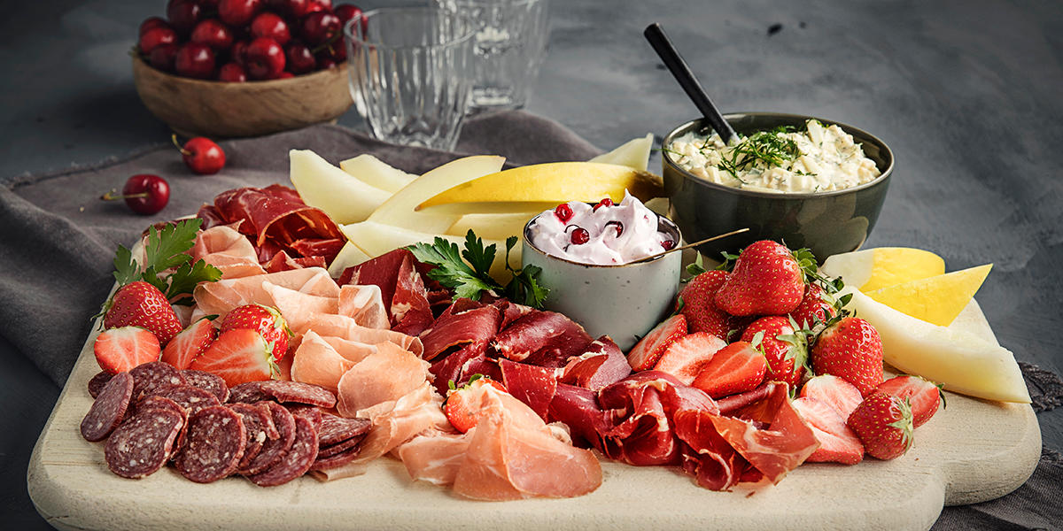 Spekefat med trollkrem og eggesalat servert på mørkt bord med en skål moreller i bakgrunnen. Spekematen er pent dandert, og eggesalaten og trollkremen er i små skåler på fatet.