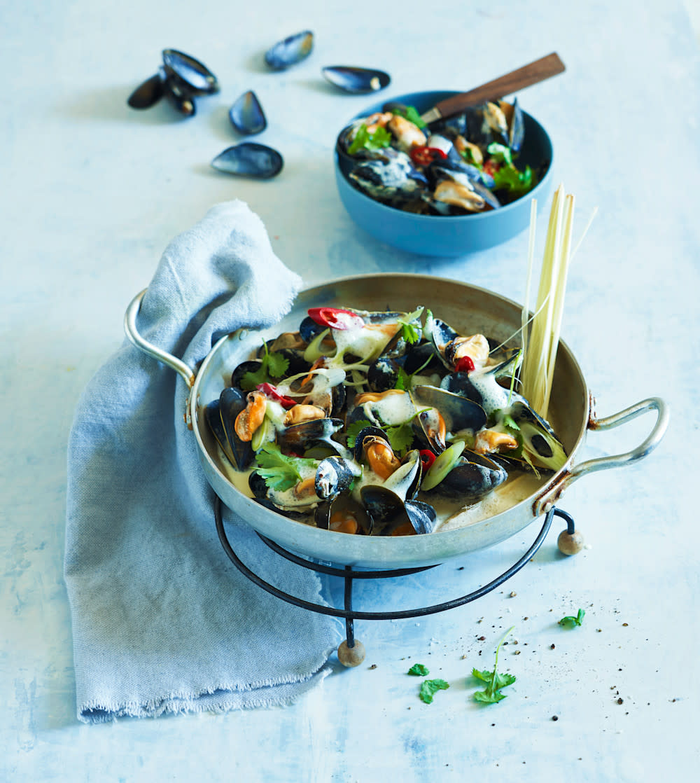 Dampede blåskjell i grønn curry saus servert i metallkjele på svart rist. Finsnittet vårløk, chili og ferske urter strødd over. Liten skål med blåskjell i bakgrunnen.