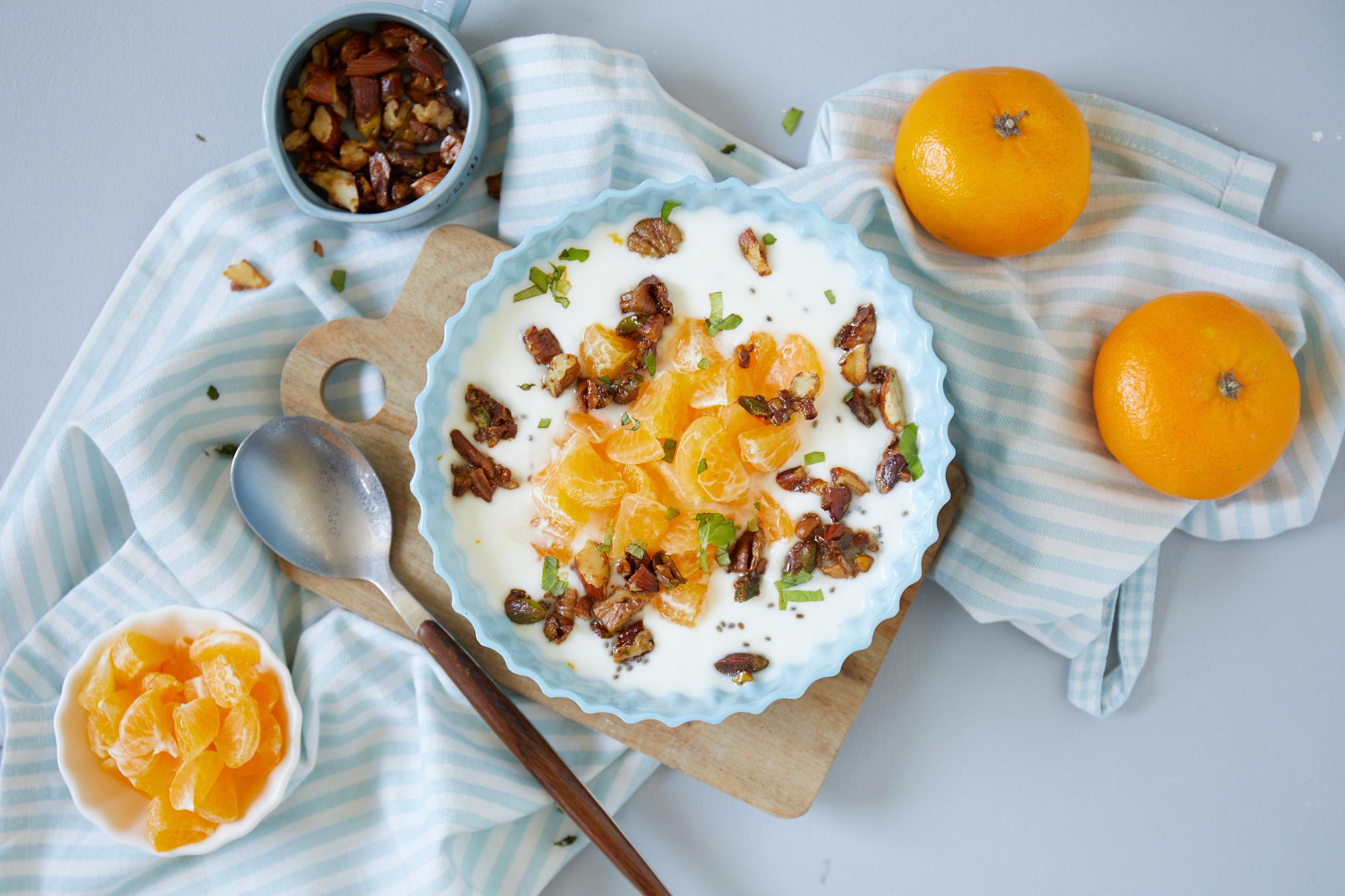 Yoghurt bowl med klementin og honningglaserte nøtter servert i lyseblå skål med et dryss frisk mynte. Hele klementiner og små skåler med klementin og honningglaserte nøtter rundt.