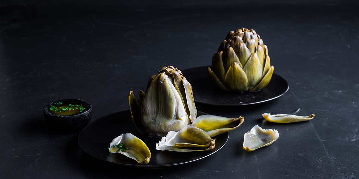 Hel artisjokk med hjemmelaget fransk dip, servert på to mørke tallerkner på mørk bakgrunn.