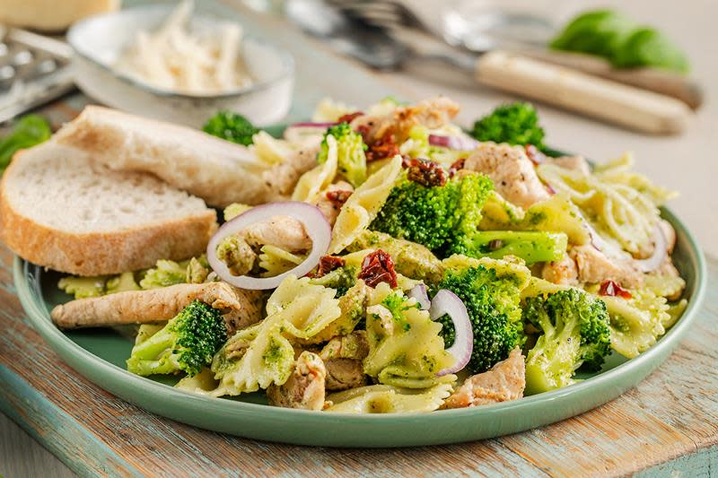 Mettende pastasalat med kylling og pesto, brokkoli og rødløk. Servert på grønt fat med lyst brød og revet ost på siden.