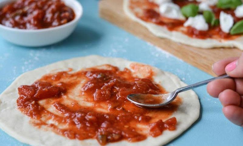 Rund pizzabunn ligger på lyst blått underlag. Hånd brer tacosalsa over bunnen.