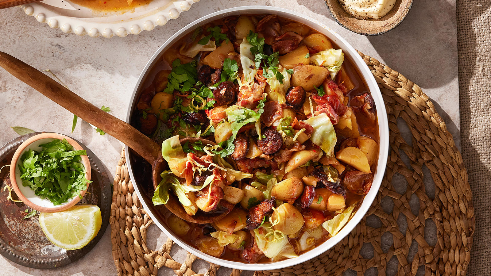 Spansk gryte med chorizo og nykål servert i hvit form med rustikk tresleiv stukket i. Limebåt og liten skål med urter ved siden av.