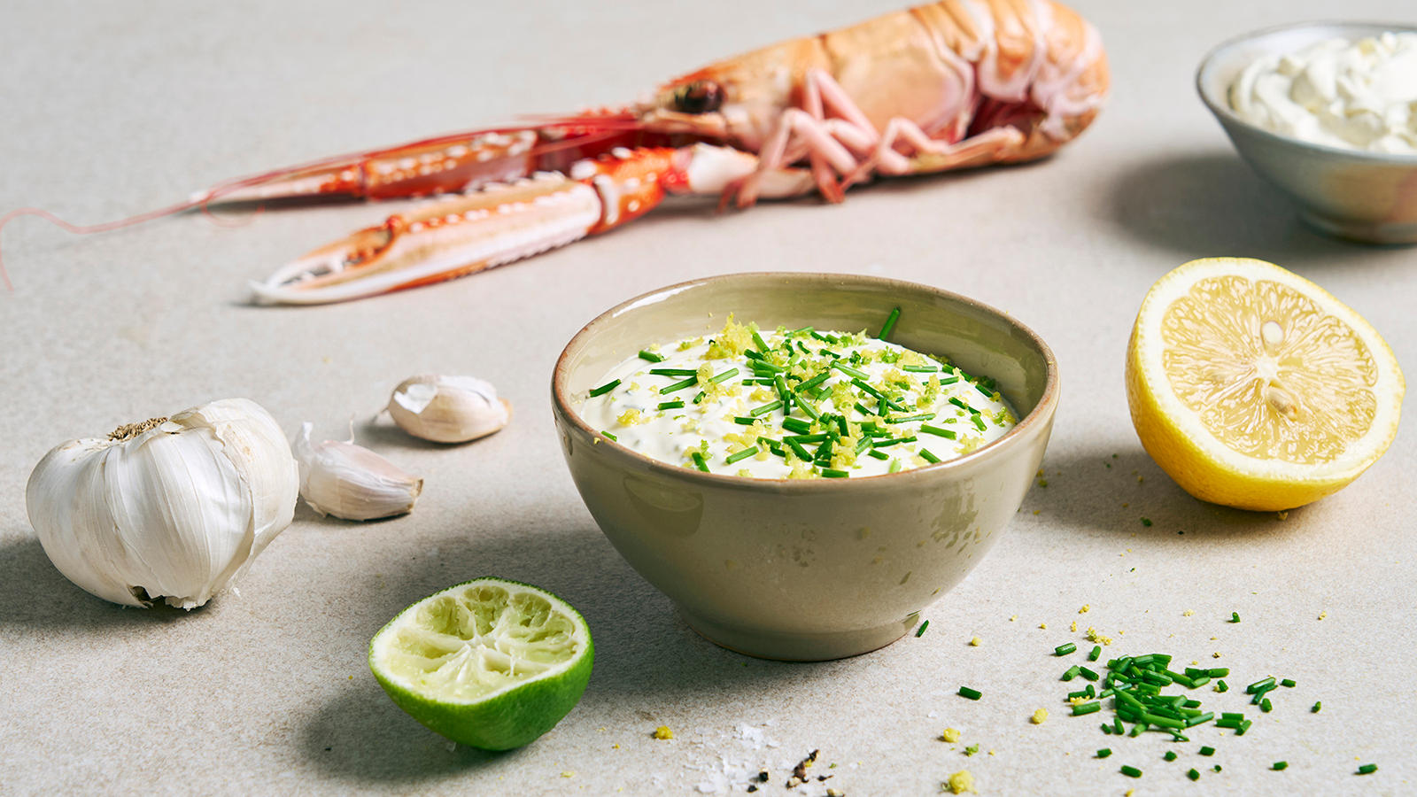 Hvitløksdressing med sitron og lime servert i liten keramikkskål med gressløk og sitronskall på toppen. Halv lime og sitron på siden, og sjøkreps i bakgrunnen.