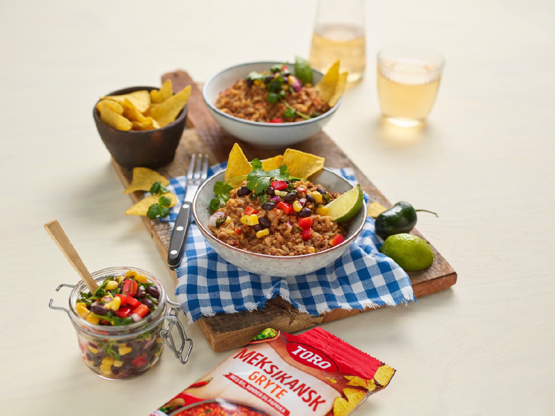 2 porsjoner meksikansk bowl på en fjøl med tortillachips og kålsalat til vestre og Toro meksikansk pose i forgrunnen samt jalapeño og lime til høyre for skålen