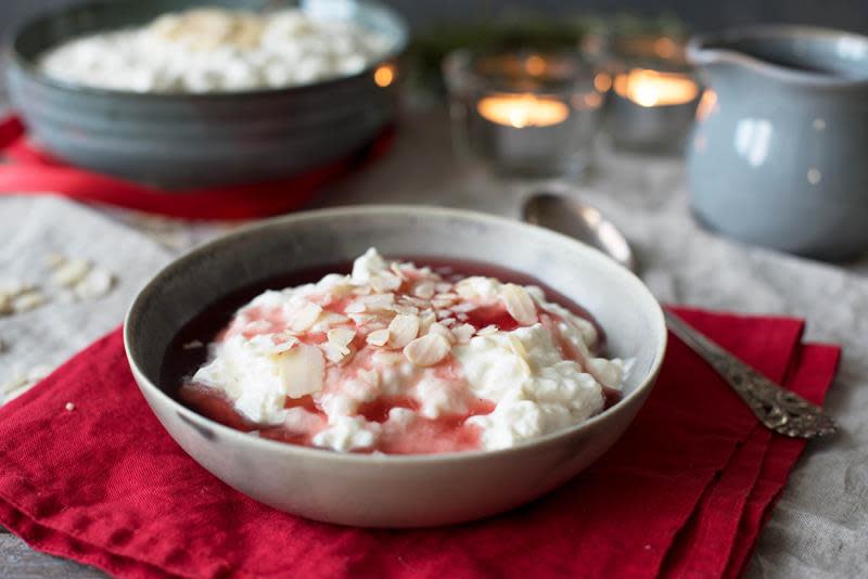 Lys skål med riskrem og rød saus, toppet med skivede mandler. Servert på rød linserviett, med telys i bakgrunnen.