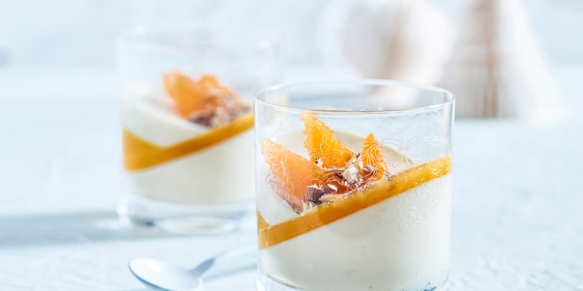 Panna cotta med hjemmelaget appelsingelé servert lagvis i to glass med appelsinfileter og hakkede nøtter på toppen. Glassene står på hvitt bord med hvit julepynt i bakgrunnen.