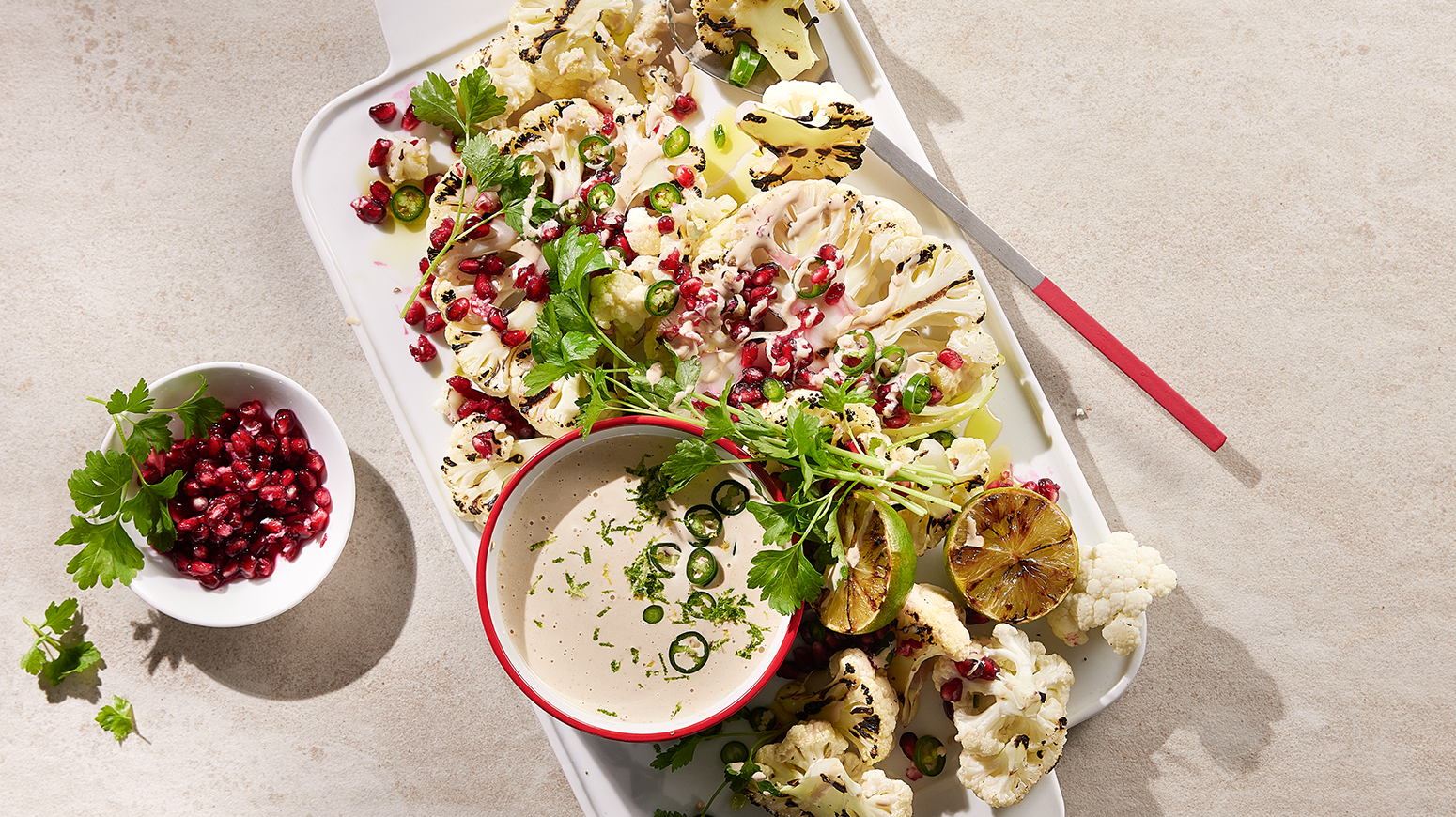 Grillet blomkål med peanøttsaus og granateple servert på en hvit tallerken med friske urter, halve lime og en skål peanøttsaus. Liten skål med granateplefrø ved siden av.