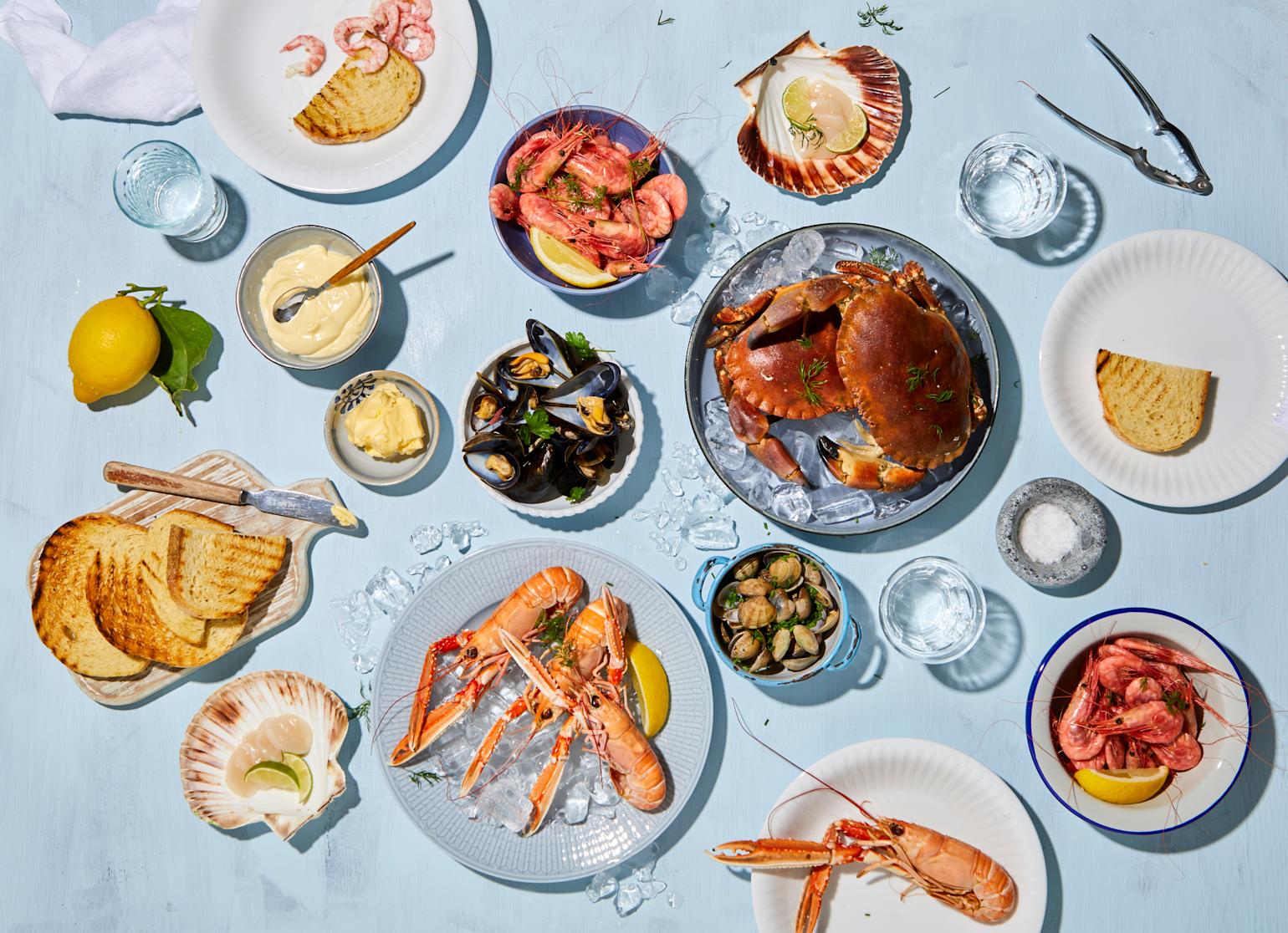 Et utvalg skalldyr på forskjellige fat og i skåler, omringet av skåler med tilbehør, ristet brød og frisk sitron.